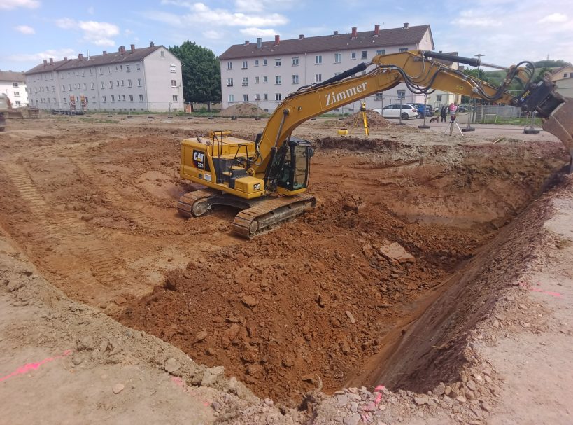 Gelbes Baggergerät gräbt in einer Baustelle umgeben von Wohngebäuden.