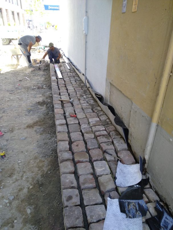 Arbeiter verlegen Pflastersteine auf einem Gehweg neben einer Hauswand.