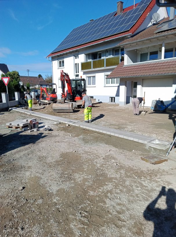 Bauarbeiten mit Bagger und Arbeiter auf einem unbefestigten Gelände neben einem Mehrfamilienhaus.