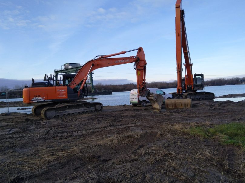 Bagger arbeiten am Ufer eines Gewässers, umgeben von Erde und Gras.