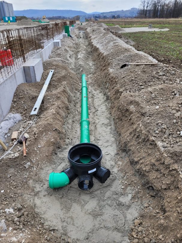 Grüne Abwasserrohre liegen in einem ausgehobenen Graben auf unbefestigtem Boden.