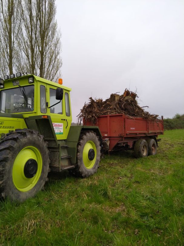Grüne landwirtschaftliche Maschine zieht einen roten Anhänger mit Pflanzenmaterial.