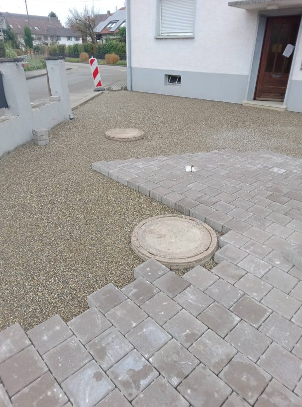 Kiesfläche mit zwei runden Kanalschächten und Pflastersteinen.