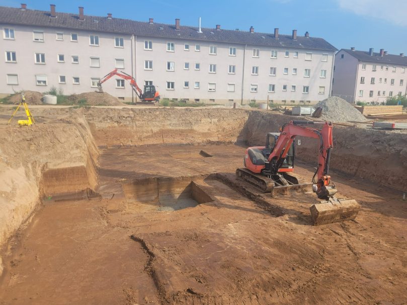 Bagger arbeitet auf einer Baustelle mit gegrabenem Boden und Wohngebäuden im Hintergrund.