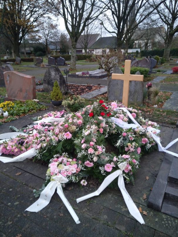 Blumengestecke mit weißen Bändern auf einem Friedhof, neben einem Kreuz aus Holz.
