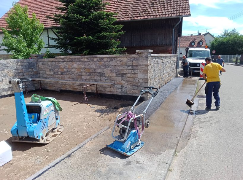 Bauarbeiter mit Maschinen und Lkw auf einer Baustelle neben einem gemauerten Zaun.