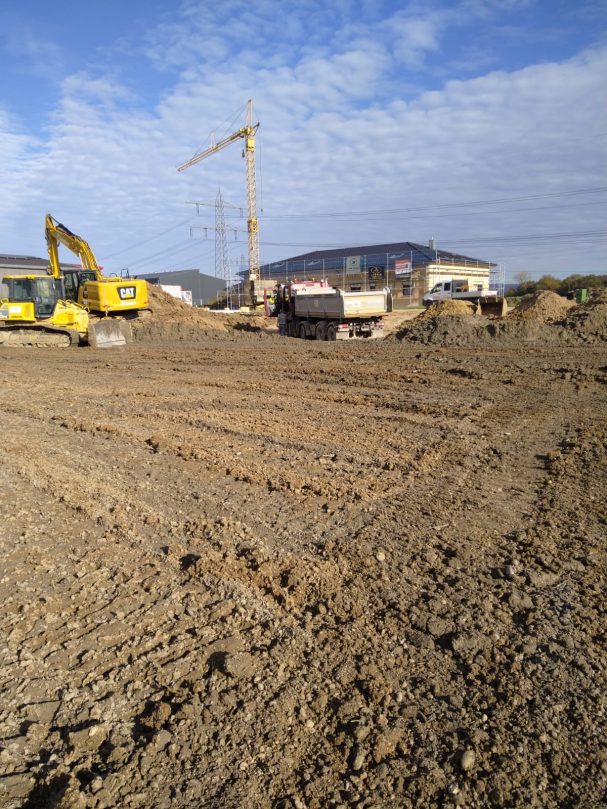 Baufeld mit schwerem Gerät, unbefestigtem Boden und Kränen im Hintergrund.