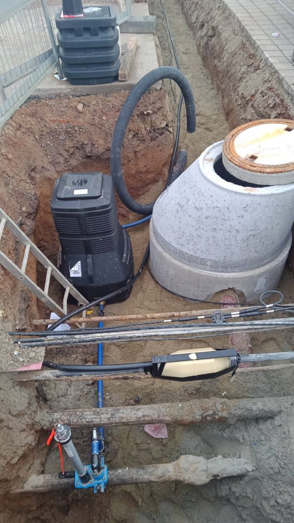 Graben mit einem Betonbehälter und einem schwarzen Gerät, Rohrleitungen und einer Leiter.